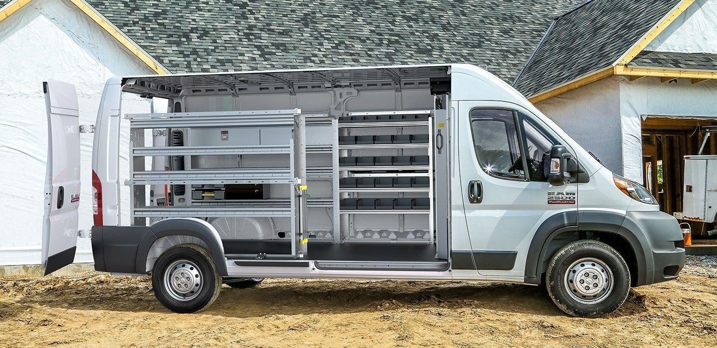 A white Ram ProMaster commercial van parked outdoors. The side sliding door is open to reveal an interior upfitted with professional metal storage shelving. A silver ladder rack is installed on the roof